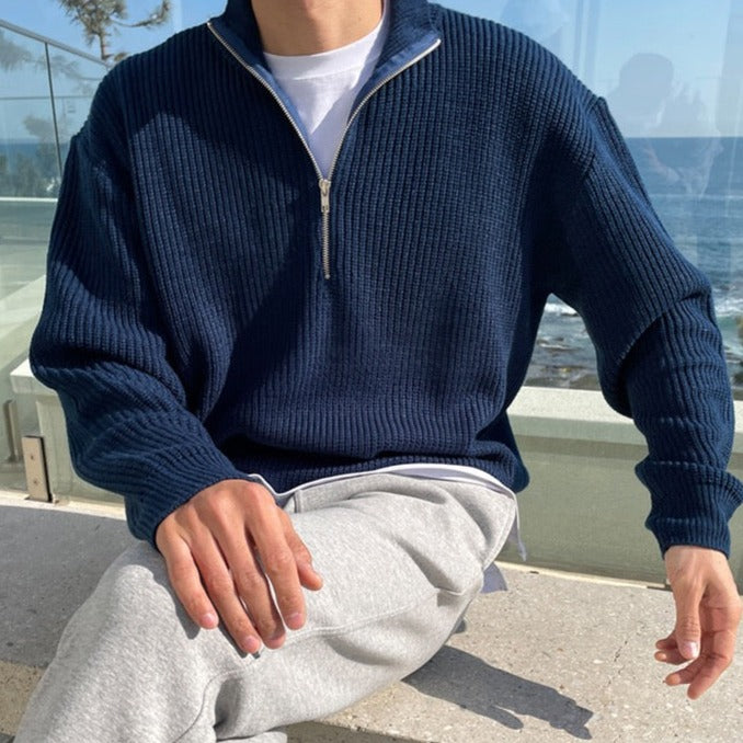 Person wearing a navy blue zip-up sweater and gray pants sitting outdoors.