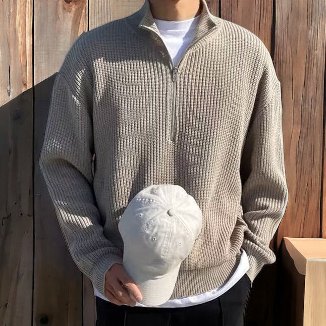 Person wearing a beige ribbed sweater holding a white cap against a wooden background