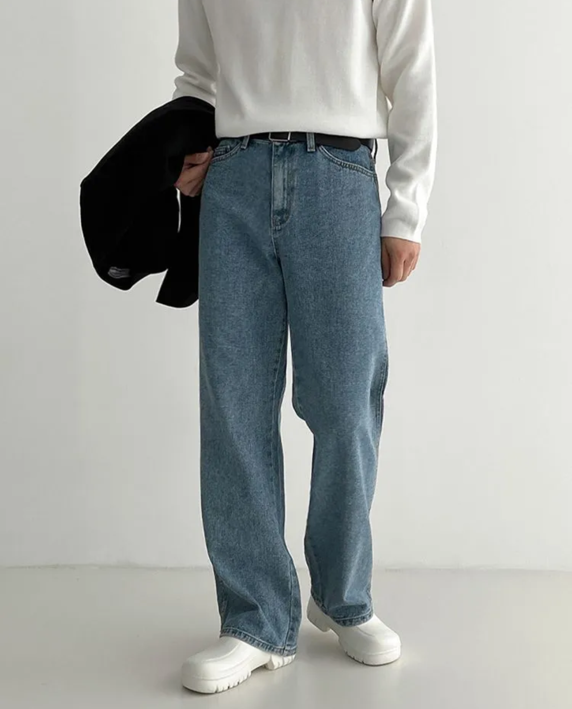 Person wearing a white long-sleeve shirt, blue jeans, and white sneakers holding a black jacket against a plain background.