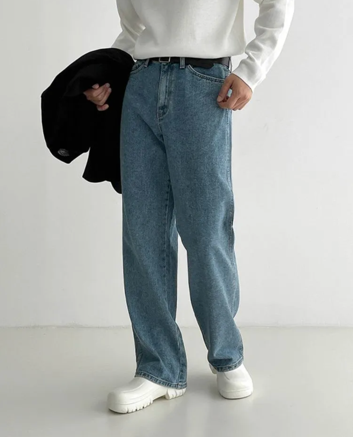 Person wearing blue jeans and a white shirt holding a black jacket against a white background