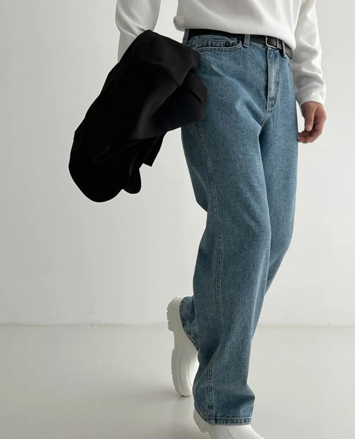 Person wearing blue jeans and white shoes holding a black jacket on a plain background