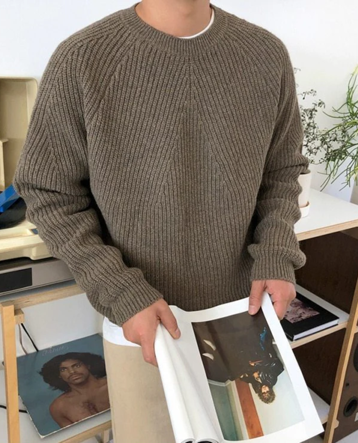 Person wearing a brown ribbed sweater holding an open magazine with a blurred face on a blurred indoor background