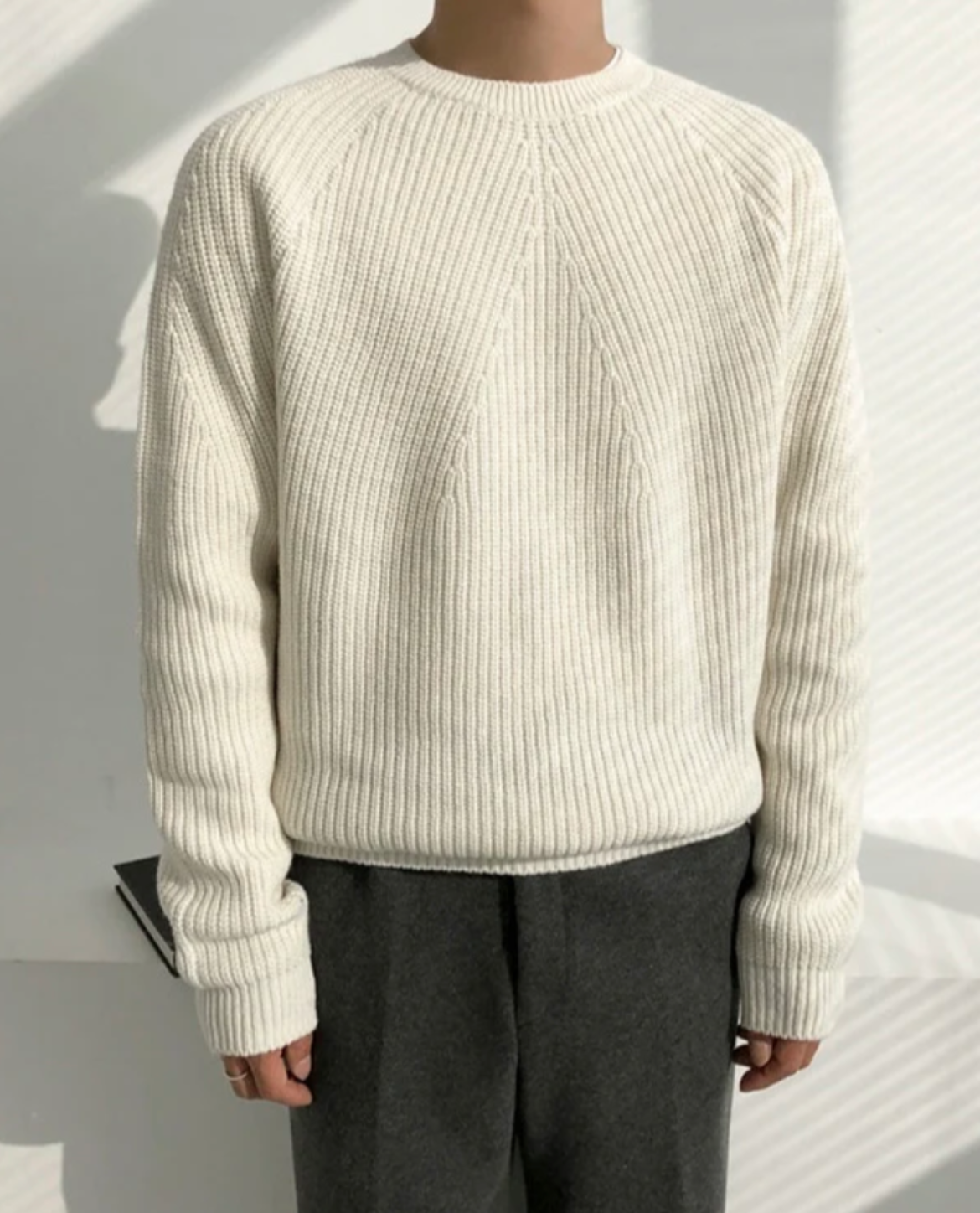 Person wearing a cream ribbed sweater and dark pants against a white background