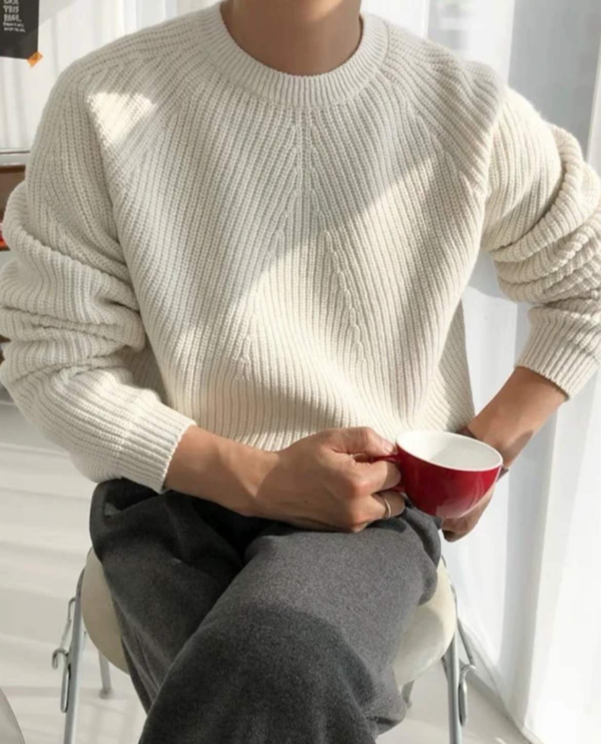 Person wearing a beige sweater and gray pants, holding a red mug indoors.