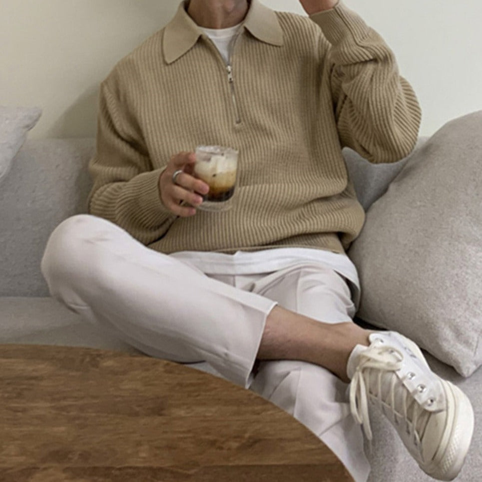 Person sitting on a couch wearing a beige sweater, white pants, and white sneakers, holding a glass of coffee.