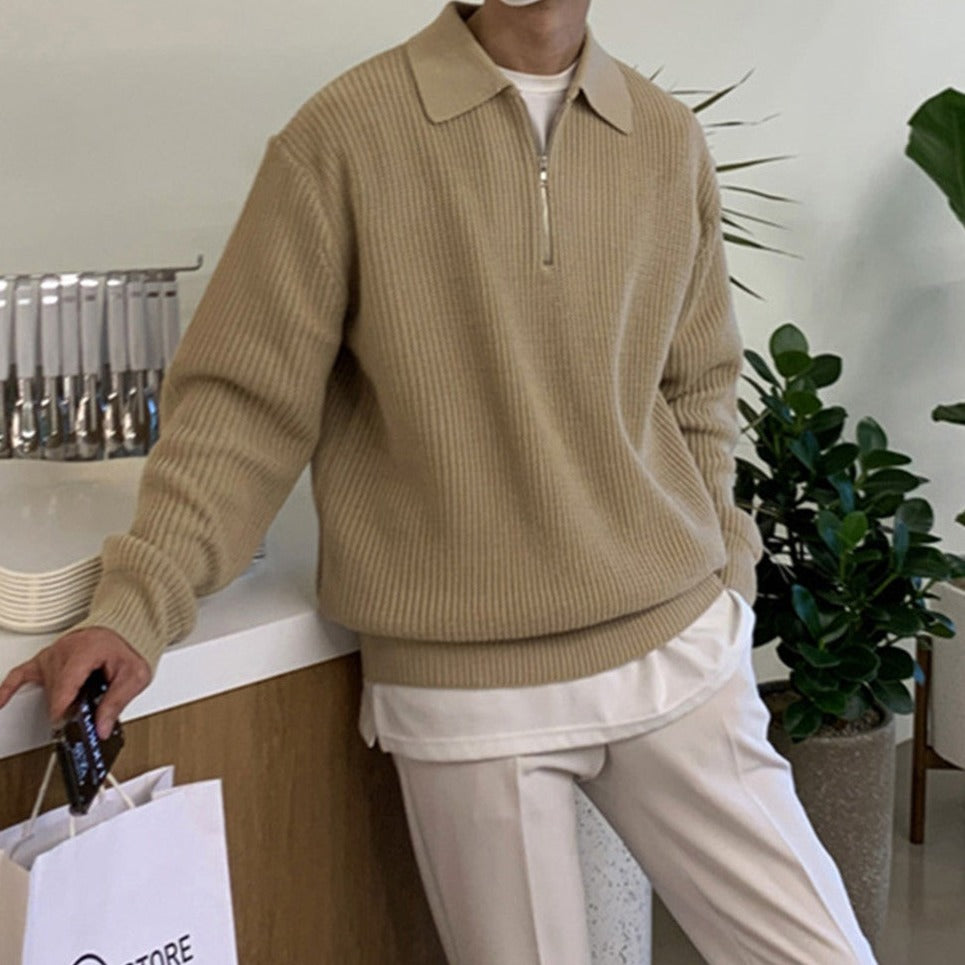 Person wearing a beige sweater and pants in an indoor setting with a plant and counter.