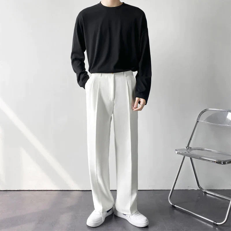 Person wearing a black sweater and white pants standing against a plain background with a chair visible.