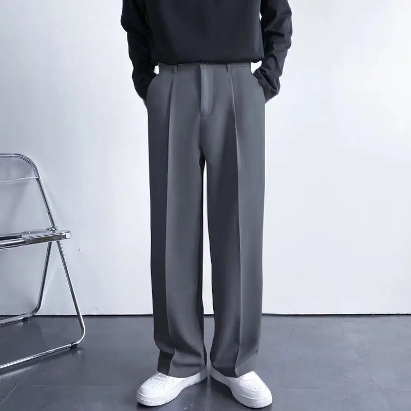 Person wearing gray pants and a black top in a minimalistic setting with a chair and white wall.
